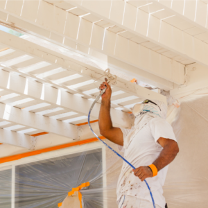 Painter spray-painting an exposed ceiling while wearing protective clothing