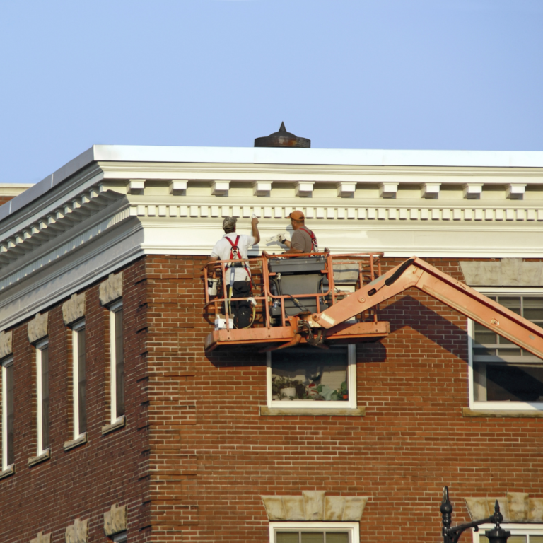 Workers on a lift beside a brick building's exterior trim